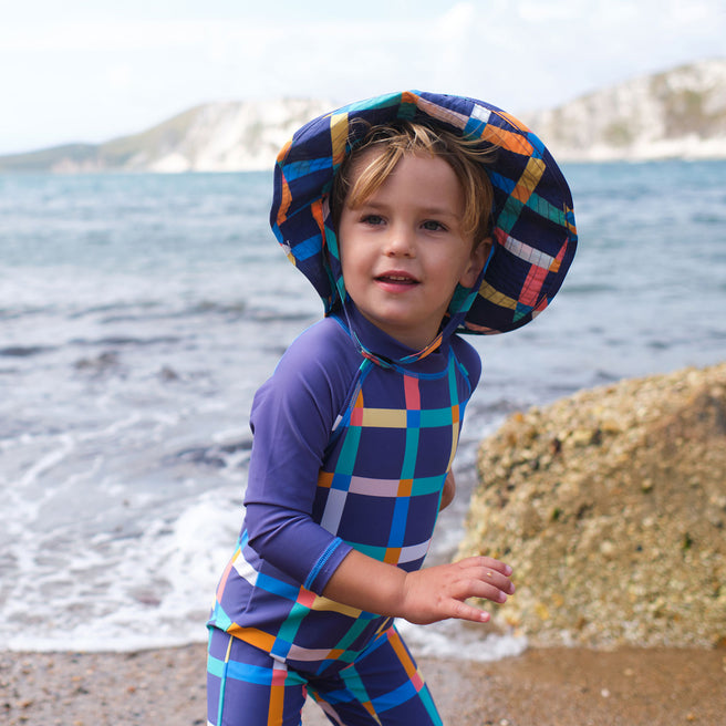 A child on a beach wearing a UV Protective Sun Hat Multi Check, in a blue checked swimsuit.